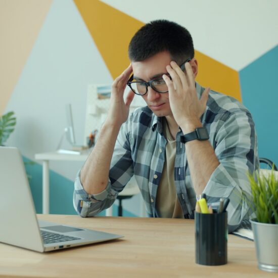 Stressed guy in glasses is working with laptop computer in office suffering from headache overworking touching head massaging temples. Work and health concept.
